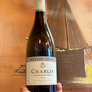 A hand holding a bottle of Bernard Demaille Chablis 2024 white wine against a warm-toned painting of a sailing ship.