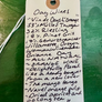 A handwritten wine tag on cream paper with a wooden grommet and white string, listing several Oay Wines, photographed against a textured green background.