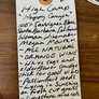 A handwritten kraft paper tag with a hole and white string, placed on a wooden surface. The tag lists ingredients and notes for a product titled "High Camp 'Happy Canyon'" in cursive handwriting.