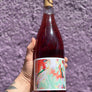 Bottle of red wine held by a person with a colorful label against a purple textured background