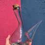 Clear glass bottle with a narrow neck and wide base held against a blue and pink wall.