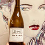 Wine bottle labeled 'ENKIDU' held in front of a stylized face artwork.