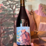 Wine bottle with a colorful label featuring a bear design, held in front of a scenic painting.