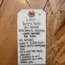 A handwritten white paper tag with a hole and string, placed on a wooden surface. The tag lists ingredients and notes for a green banana bread recipe in handwritten black ink.