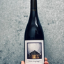 A hand holds a dark glass wine bottle with a white label featuring a black-and-white architectural photograph and the handwritten text 'Woman, diest fragte?'. The background is a textured gray wall.