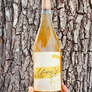 A hand holds a bottle of MianU wine against a textured tree trunk background. The bottle contains a golden-amber liquid and has a cream label with a yellow brushstroke design and the brand name 'Niman' in black script.
