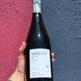 A hand holding a bottle of Mirgin Reserva 2021 red wine, set against a split pink and dark blue textured wall.
