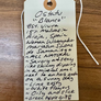 A handmade paper hangtag with handwritten Spanish text describing a natural olive body wash, tied with a white string and photographed on a wooden surface.