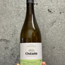 A hand holding a bottle of Ostatu Rioja Alavesa Blanco 2020 white wine against a textured gray concrete wall.
