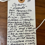 A handwritten white paper price tag with a wooden dowel hole and string, showing recipe details for 'Ostatu Rosado' jam on a wooden surface.