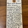 A handwritten cream-colored paper tag with a brown circular hole at the top, featuring cursive writing about 'Poggio delle Basciati' wine, placed on a wooden surface.