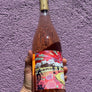 A hand holding a wine bottle with a dark brown wood-grain style decanter body and a colorful abstract label, set against a textured purple wall.