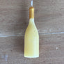 A novelty bottle-shaped candle in pale yellow wax with a metallic gold neck and a white wick, placed on a brown wooden surface.