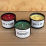 Three black soy candles in round black jars on a wooden surface, labeled Sapphoir Faire: VIVA LA VULVA, ABUNDANCE, and QUEER JOY, with red, green, and yellow wax visible.