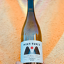 Wine bottle labeled 'Multitudes' held against a textured orange background
