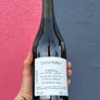 A hand with a floral tattoo holds a dark glass bottle of wine against a split blue and pink textured background. The bottle has a white label with handwritten text.