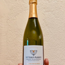 A hand holding a bottle of Vitteaut-Alberti Cremant de Bourgogne Blanc de Blancs sparkling wine against a textured beige wall.