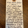A handwritten wine tag for Alta Alella Cellar 'AVS' Pet-Nat Red, detailing the wine's characteristics and ingredients, placed on a dark wooden surface.