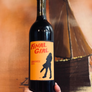 A hand holds a dark wine bottle with a bright orange label that reads 'FINAL GIRL' and 'SANGIOVESE 2024' against a warm brown background with a ship illustration.
