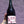 A hand with a floral tattoo holds a large dark glass bottle of Folicello Il Rosato Rosato Valti wine against a textured purple stucco wall.