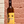 Wine bottle with a yellow label featuring a face design, held against a brick wall.