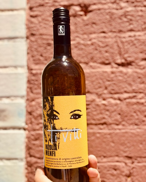 Wine bottle with a yellow label featuring a face design, held against a brick wall.