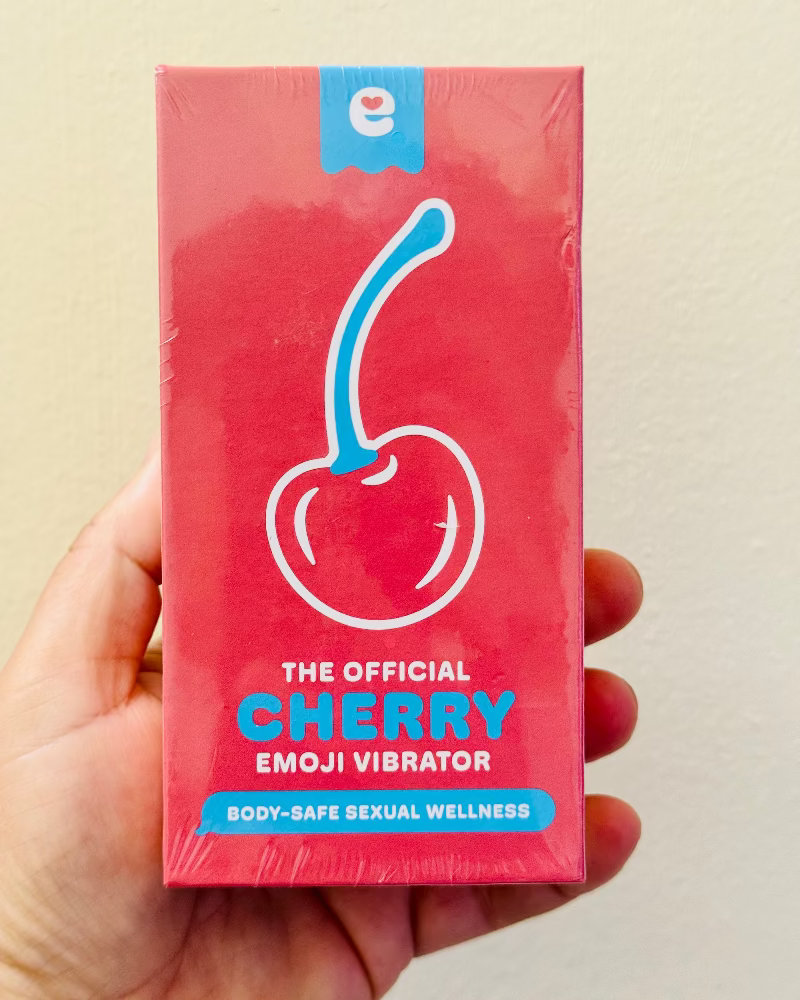 Hand holding a red packaging of 'The Official Cherry Emoji Vibrator' against a beige background
