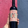 Wine bottle held by a hand with a colorful label against a pink and blue background