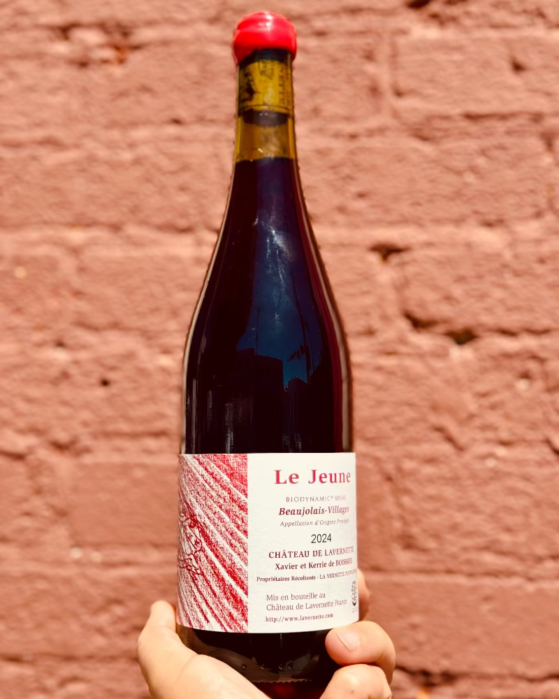 Bottle of red wine labeled 'Le Jeune' held against a brick wall.