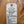 A handwritten white paper tag with a hole and string, placed on a wooden surface. The tag lists ingredients and notes for a green banana bread recipe in handwritten black ink.