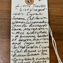 A handwritten gift tag on a wooden surface, reading 'Little Trouble "Gist Vineyard" 100% Syrah Sonoma, California Women Winemakers Jennifer Reichhardt Sara Morgenstern ~ All NATURAL ~ A brimming basket of boysen, marion and cosan berries ~ Salted Swedish licorice ~ Cali Rhone vibes ~ Russian Curant tea ~ Spicy black currants ~ Freshness suprise with rich pop !$'.