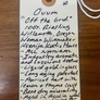 A handwritten cream-colored paper tag with a wooden punch hole and white string, printed with the text 'Ovum "Off the Grid" 100. Riesling Willamette, Oregon Woman Winemaker Ksenija Kostiā House ~ All Nature ~ Intensey aromatic, and luscious textured ~ Liquid gold in glass ~ Long aging potential ~ Apricot tart with lemon + thyme crust ~ Bone dry minerality ~ Aged in Acacia wood ~ Like zip lining for your mouth' on a dark wood background.