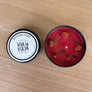 An open round black tin of red wax candle with visible chunks, next to its white lid labeled 'SAPPOIR FAIRE VIVA la VULVA LUSTY VIBES' on a light wood surface.