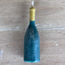 A small bottle-shaped candle in a dark teal blue color with a gold-colored neck and a white wick, lying horizontally on a light brown wooden surface.