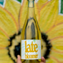 A hand holding a bottle of Late Bloomer sparkling wine with a vibrant yellow background featuring sunflower designs.