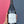A hand with a floral tattoo holds a dark glass bottle of wine against a split blue and pink textured background. The bottle has a white label with handwritten text.