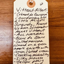 A handwritten cream-colored paper tag with a wooden tab at the top, reading 'Vitteau-Albert' and describing a Chardonnay and Aligotte Burgundy wine blend, photographed on a dark wood surface.