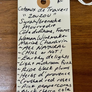 A handwritten cream-colored paper wine tag with a wooden hanging hole, placed on a brown wooden surface. The tag lists wine cellar details and flavor notes for a wine in Coteaux de Travers 'ZOUZOU' with varietals like Syrah, Grenache, and Mouvredhe, and notes 'ALL NATURAL' and 'CHILL OR NOT!'.