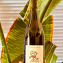 A hand holds a bottle of Zouzou wine against a background of large green tropical leaves and white window blinds.